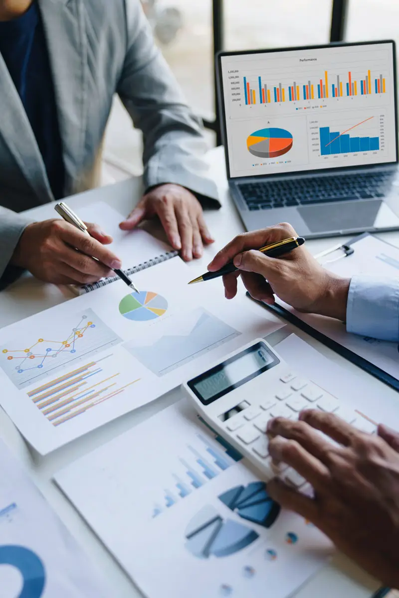 Consultants confer over a desk covered with graphs. One consultant holds a pen and crunches numbers on a calculator, while the other holds a pen, ready to annotate. The scene reflects collaboration and data-driven decision-making.